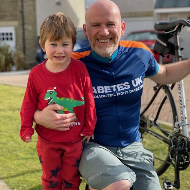 Euan in Diabetes UK cycling jersey holding his bike with his other arm around his son Archie in red pyjamas